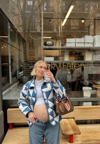 Vatteret blå og hvid jakke med geometriske mønstre, parret med en tætsiddende hvid skjorte og lyse denimjeans. Holder et glas og bærer en brun taske.