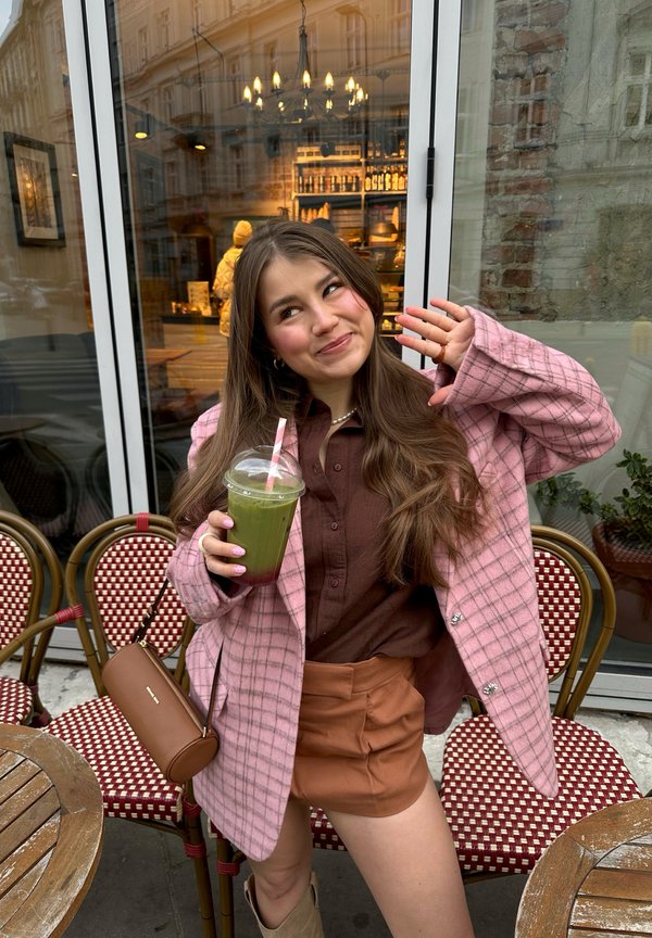 Ung kvinde i lyserød ternet jakke og brunt outfit holder en grøn smoothie med sugerør, smiler og vinker, mens hun sidder på en udendørs café.