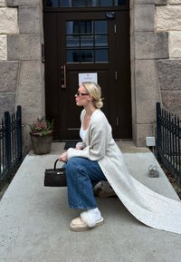 Femme accroupie sur le trottoir portant un long cardigan blanc, un jean bleu, des mocassins doublés de fourrure, des lunettes de soleil noires, tenant un sac à main noir près d'une porte en bois sombre.