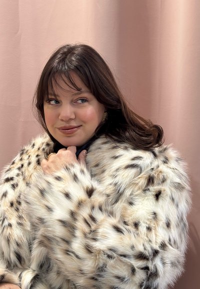 Joven con cabello largo y castaño, vestida con un abrigo de piel sintética blanca con manchas, mirando de lado con una suave sonrisa sobre un fondo rosa claro.
