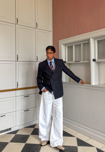 Hombre de pelo oscuro lleva una americana de rayas, camisa a rayas, corbata y pantalones blancos anchos, de pie en una cocina con armarios blancos.