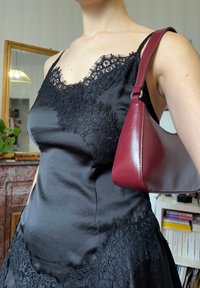 Femme portant une robe en satin noire bordée de dentelle et un sac à main bordeaux brillant sur l'épaule, à l'intérieur avec un miroir et des livres en arrière-plan.