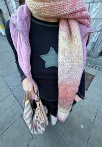 Veste matelassée rose, chemise noire avec écusson étoile verte, écharpe tricotée rose-jaune, sac rayé aux tons crème et marron, et baskets claires.