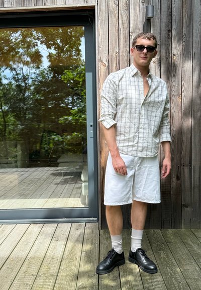 Camisa blanca de cuadros, pantalones cortos blancos, zapatos negros y calcetines blancos, con un fondo de una terraza de madera y una puerta de cristal donde se puede ver vegetación.