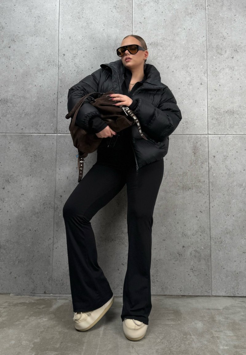Chaqueta tipo puffer negra con cuello alto, top negro ajustado, pantalones negros de campana y zapatos de color crema. Los accesorios incluyen un bolso marrón y gafas de sol de gran tamaño.