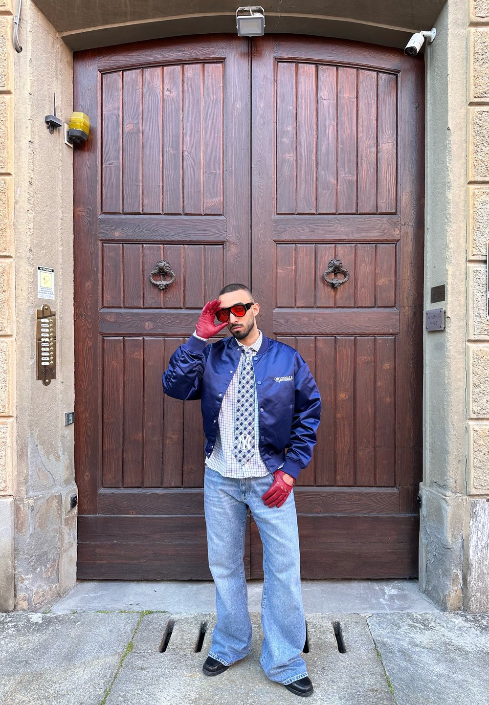 Giacca bomber in satin blu navy, camicia a quadri, cravatta con motivo, guanti rossi, jeans in denim svasati e occhiali da sole davanti a una porta di legno.