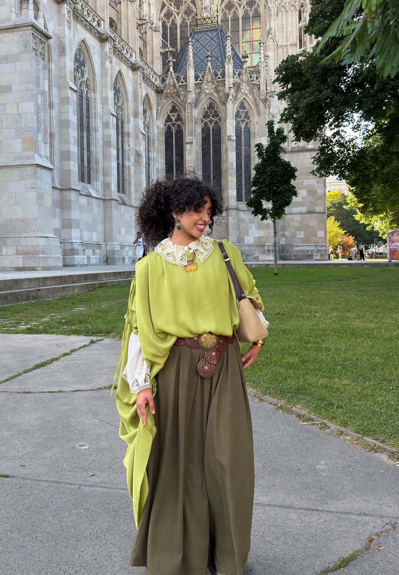 Vrouw in groene blouse en lange bruine rok staat bij een gotisch stenen gebouw met grote glas-in-loodramen en nabijgelegen groene bomen.