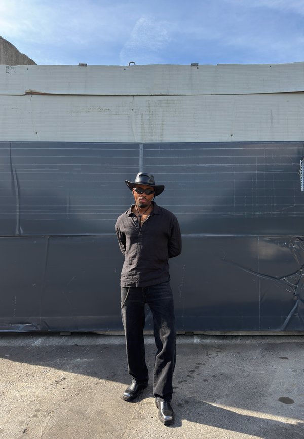 Homme portant chemise noire, pantalon, bottes, chapeau à large bord et lunettes de soleil, debout les mains dans le dos devant un mur industriel gris.
