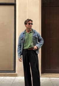 Young man wearing sunglasses, a green shirt, a denim jacket, black trousers, and rings stands against a beige wall near a wooden door.