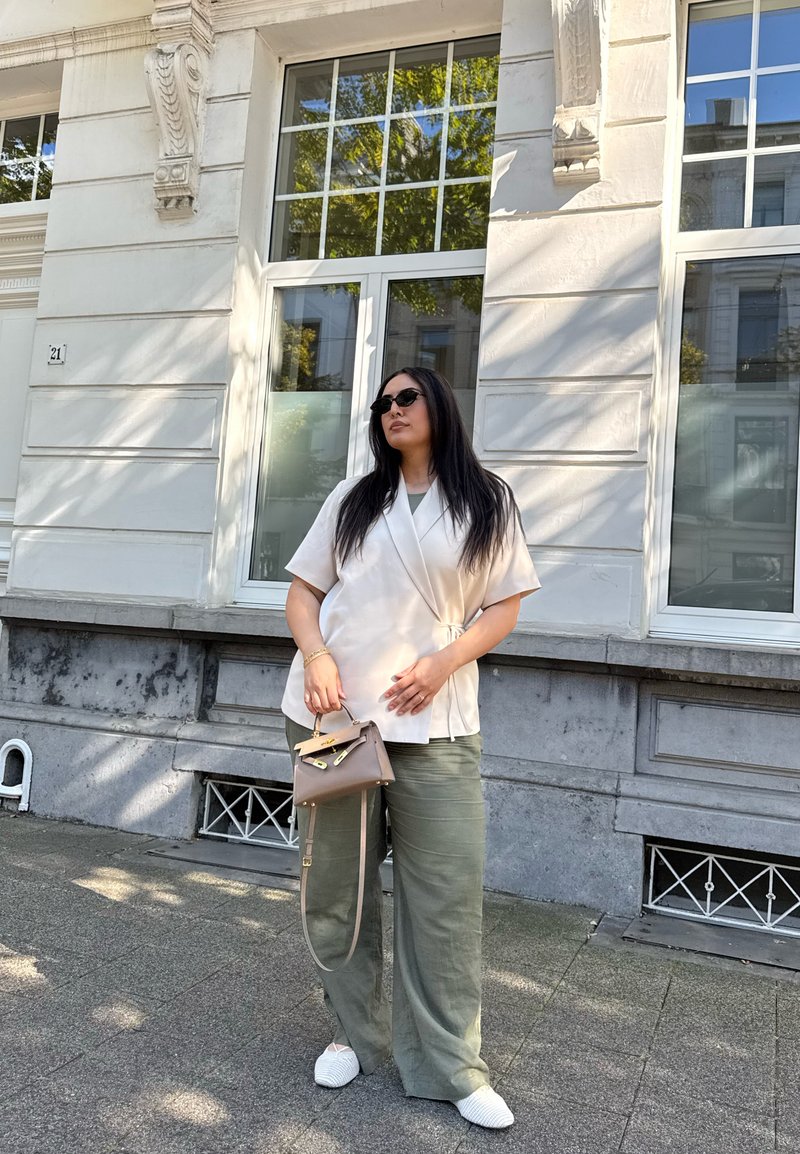 Blusa envolvente bege, calças largas verde-oliva, sapatos brancos de slip-on texturizados e uma pequena mala castanha com alça para o ombro.