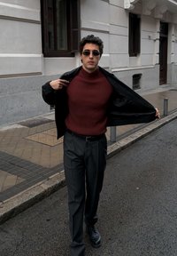 Νεαρός άνδρας με γυαλιά ηλίου, μπορντό πουλόβερ και μαύρο παντελόνι περπατά σε δρόμο της πόλης, ανοίγοντας το μαύρο σακάκι του για να αποκαλύψει την εμφάνισή του.