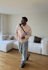 Homme en veste beige et jeans bleu clair tenant un téléphone, debout dans un salon avec un canapé sectionnel blanc et un sol en bois.