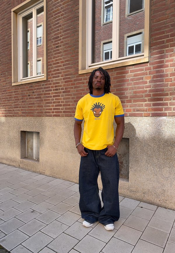 Homme avec des dreadlocks portant un tee-shirt jaune avec un motif graphique et un jean large et foncé, debout sur un trottoir carrelé près d’un mur en briques avec des fenêtres.