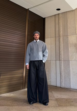 Hombre con sombrero gris, chaqueta gris corta, camisa blanca, corbata negra y pantalones anchos negros, de pie junto a una puerta marrón y una pared beige.