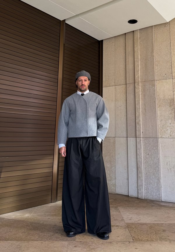 Homme portant un chapeau gris, une veste courte grise, une chemise blanche, une cravate noire et un pantalon large noir, debout près d'une porte marron et d'un mur beige.