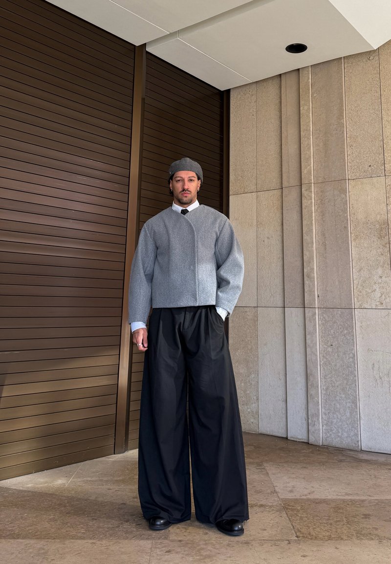 Άνδρας που φοράει γκρι καπέλο, κοντό γκρι σακάκι, λευκό πουκάμισο, μαύρη γραβάτα και φαρδιά μαύρα παντελόνια, στέκεται δίπλα σε καφέ πόρτα και μπεζ τοίχο.