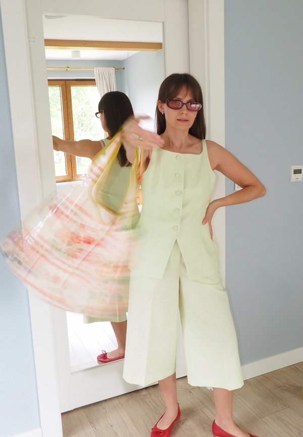 Vrouw in lichtgroene mouwloze outfit en rode schoenen gooit een kleurrijke sjaal in een kamer met een spiegel van volle lengte en houten vloer.