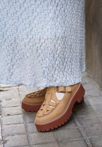 Ταμπά παπούτσια πλατφόρμας με λουράκια με κοψίματα και πόρπες, φορεμένα με λευκή υφασμάτινη φούστα πάνω σε γκρι πλακόστρωτα.