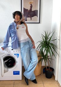 Blårandig skjorta med blommönster, vit tanktop, ljusblå baggyjeans och svarta spetsiga skor; tvättmaskin och krukväxt i närheten.