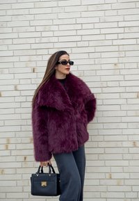 Bordo umetno krzneni plašč z širokim ovratnikom, nošen čez črno majico in temne hlače. Črna torbica z zlatim detajlom ključavnice.