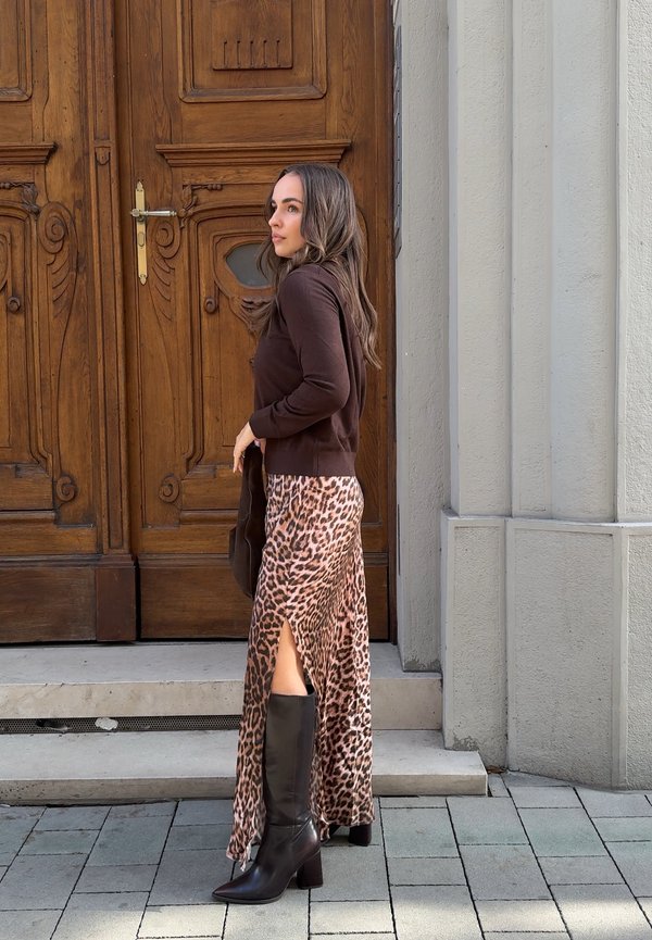 Mujer con pelo largo y castaño que lleva un suéter oscuro, falda con estampado de leopardo y abertura lateral, y botas negras hasta la rodilla, de pie junto a una puerta de madera.