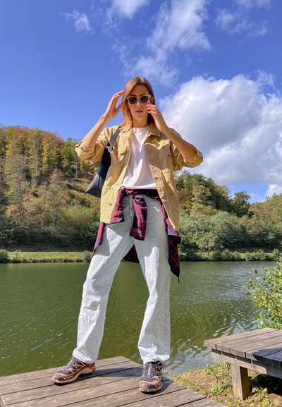 Camisa de lona beige, camiseta blanca, camisa de cuadros atada a la cintura, vaqueros azul claros, zapatos de senderismo marrones, de pie junto al agua con árboles al fondo.