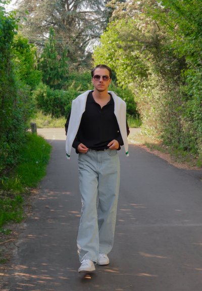 Una persona que lleva una camisa polo negra y unos vaqueros de pierna ancha de color azul claro, con un suéter blanco colgado sobre sus hombros, caminando por un sendero.