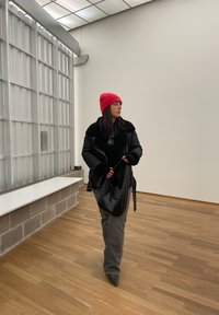 Μαύρο μπουφάν με γούνα στον γιακά, κόκκινο πλεκτό σκούφο, γκρι παντελόνι και μαύρη τσάντα. Ξύλινο δάπεδο και μίνιμαλ φόντο.