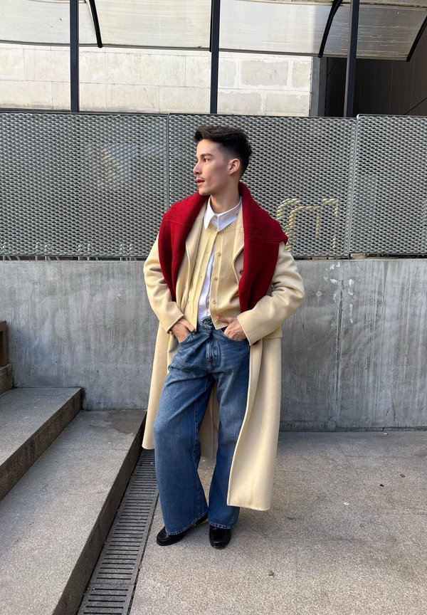 Άτομο που στέκεται έξω φορώντας μακρύ μπεζ παλτό, κόκκινο κασκόλ που πέφτει στους ώμους, κρεμ πουλόβερ, λευκό πουκάμισο και μπλε τζιν.
