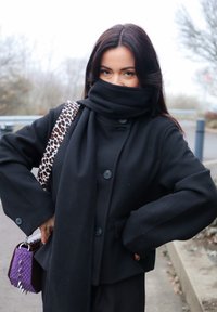 Svart ullkåpe med store knapper, kombinert med et svart skjerf. Har en lilla og teksturert veske med pigger og leopardmønstrede stropper.