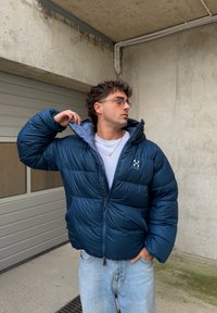 Tengerészkék steppelt dzseki magas gallérral, cipzáras záródással és logó részlettel. Fehér póló fölött viselve, világoskék farmerrel. Texturált anyag.