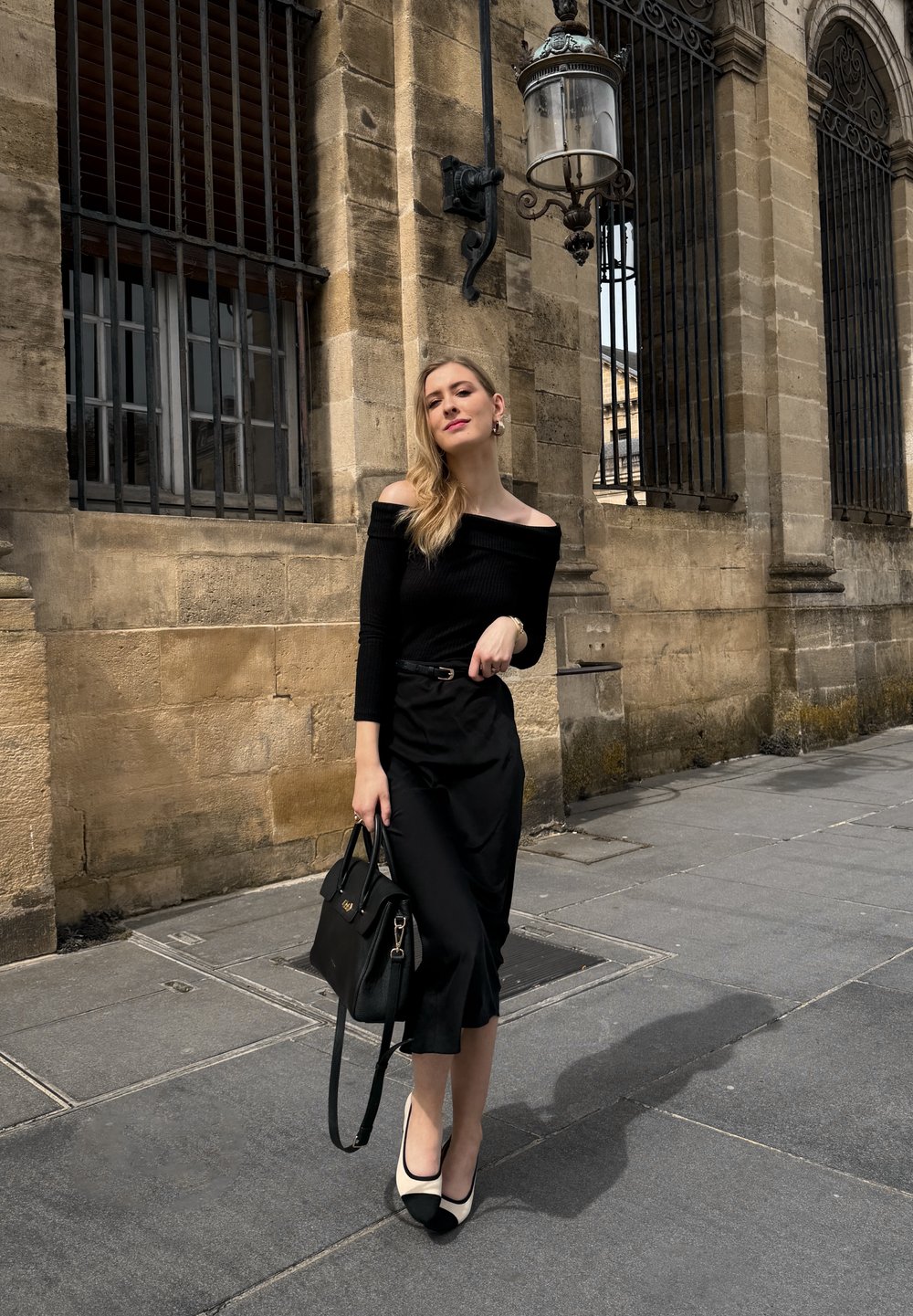 Top nero a spalla scoperta in maglia abbinato a una gonna a portafoglio nera. Gli accessori includono una borsa nera strutturata e tacchi a punta bicolore.