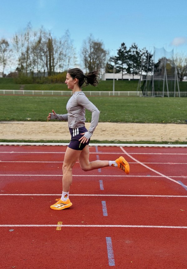 Femme courant sur une piste extérieure rouge portant un t-shirt gris à manches longues, un short violet, des chaussettes blanches et des chaussures de course orange vif.
