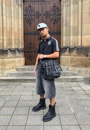 Camisa polo negra con rayas grises, pantalones cortos grises desgastados, botas negras y un bolso bandolera negro con herrajes dorados. El fondo presenta una puerta de madera.