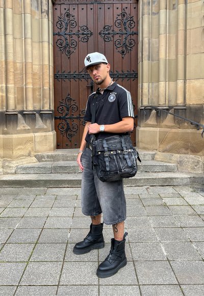 Camisa polo negra con rayas grises, pantalones cortos grises desgastados, botas negras y un bolso bandolera negro con herrajes dorados. El fondo presenta una puerta de madera.