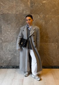 Jeune femme debout contre un mur en marbre, portant un long manteau gris, un pull gris, un pantalon blanc, des baskets, et portant un sac noir en bandoulière.