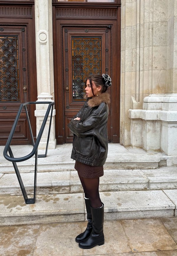 Chaqueta de cuero marrón con cuello de piel, falda color chocolate, medias negras y botas negras hasta la rodilla. Barandilla metálica y puertas de madera ornamentadas en el fondo.