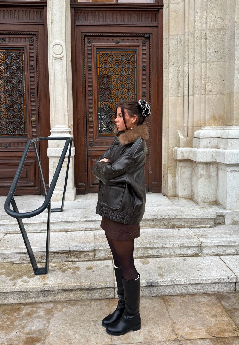 Veste en cuir marron avec col en fourrure, jupe chocolat, collants noirs et bottes noires montantes jusqu'aux genoux. Rambarde en métal et portes en bois sculpté en arrière-plan.