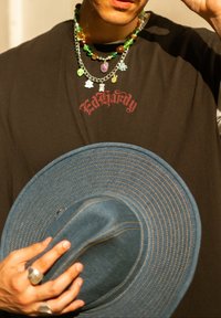 Dunkles T-Shirt mit "Ed Hardy"-Logo, geschichtet mit bunten Perlenketten. Breite blaue Hut mit kontrastierenden Nähten in der Hand gehalten.