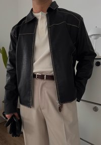 Chaqueta de cuero sintético negra con costuras en contraste, cierre de cremallera, llevada sobre una camiseta henley de canalé beige y pantalones beige, sosteniendo guantes negros.