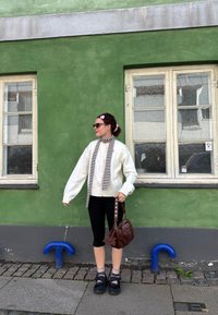 Balta per didelė megztinis su juodai baltu dryžuotu šaliku, juodos leginsai ir ruda rankinė. Stovi priešais žalią sieną su langais.
