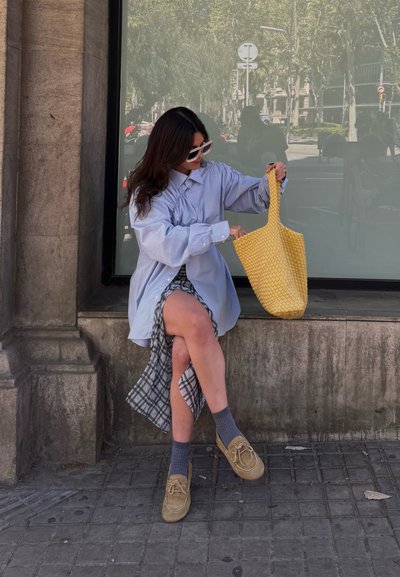 Camisa de botones oversized azul claro, falda de cuadros, bolsa de tela tejida amarilla, calcetas grises y zapatos marrones, sentado en un borde de piedra.