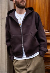 Sweat à capuche marron à fermeture éclair avec une grande capuche, des poches à l'avant et une coupe décontractée, porté sur un t-shirt blanc et un pantalon beige.