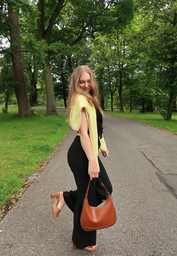 Geel gebreide cardigan, zwarte gerimpelde top, zwarte aansluitende broek, tan blokhak schoenen en een bruine schoudertas, op een met bomen omzoomd pad.