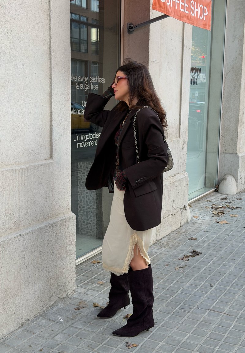 Femme portant des lunettes de soleil, un blazer noir, une jupe crème et des bottes noires, regardant la vitrine d'un café sur un trottoir en ville.