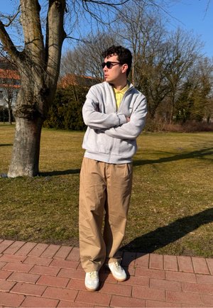 Sudadera gris con cremallera, camisa polo amarilla, pantalones beige de pierna ancha y zapatillas blancas. De pie al aire libre con un fondo de árboles y cielo azul.