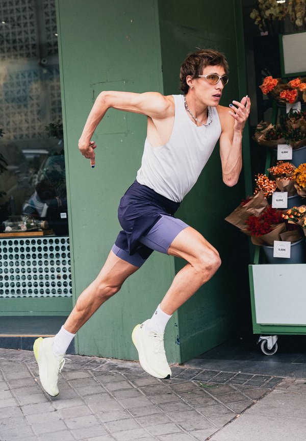 En løber i hvidt tanktop, lilla shorts og gule sko løber forbi en blomsterbod med farverige buketter udenfor en grøn butik.