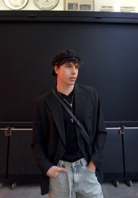 Blazer nero doppiopetto sopra un maglione nero, jeans chiari e un cappello piatto. Collane d'argento con croci; borsa a tracolla nera.