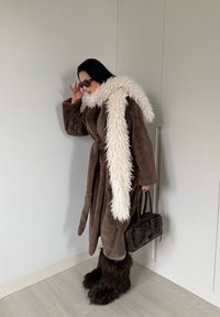 Manteau en peluche marron avec un long col en fausse fourrure blanche, couvre-jambes marron duveteux, et un sac à main noir. Texture lisse et manches larges.