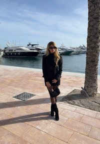 Blazer preto sobre um top justo e canelado, combinado com uma mala preta. Apresenta cabelos longos e ondulados, óculos de sol e botas pretas até ao joelho. Caminho de mármore.